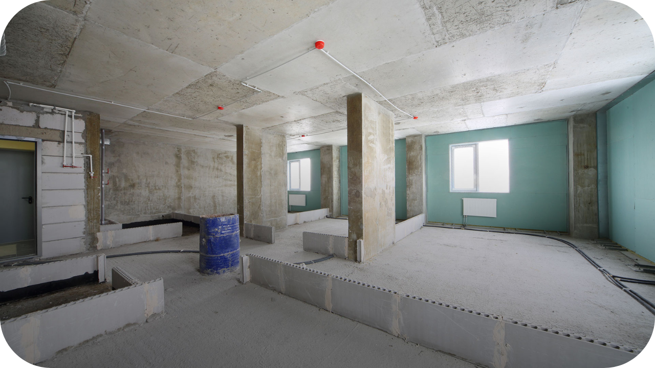 Unfinished commercial space under construction with exposed concrete walls, ceiling beams, and utility pipes installed.