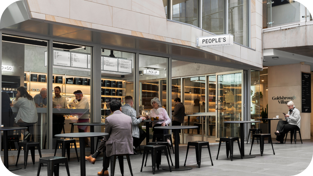 An outdoor café called PEOPLE’S, located in a shopping precinct, with customers sitting at black tables and others ordering inside through the glass storefront.