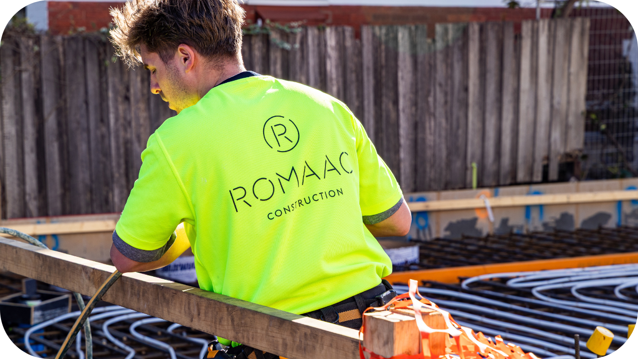 A construction worker wearing a bright yellow high-visibility shirt with the ROMAAC Construction logo on the back, working on a building foundation site with visible wiring and timber framing.