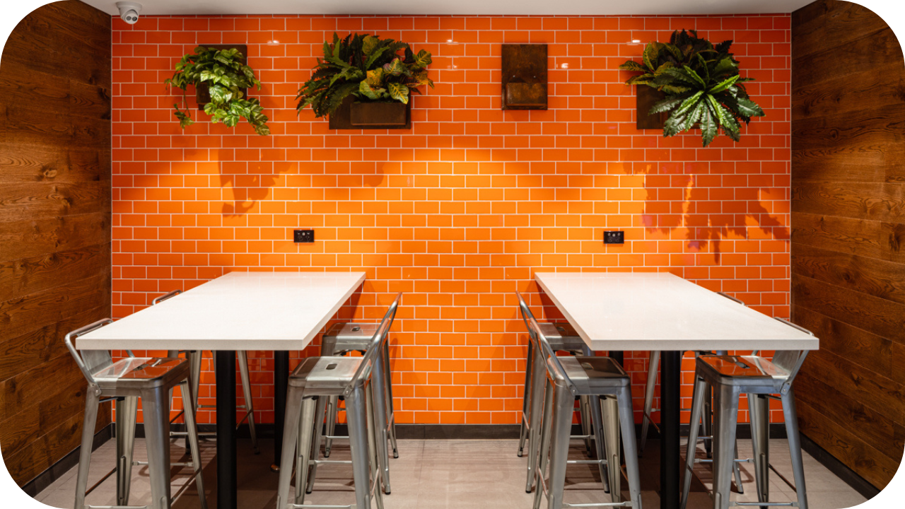 A modern break area featuring two high tables with metal stools, orange subway-tiled walls, wooden side panels, and hanging green plants for decoration.