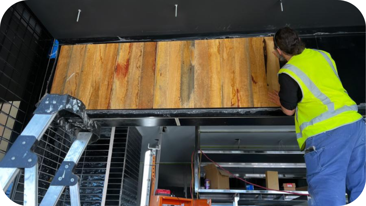 Tradesperson installing vertical timber panels under a black ceiling using plywood backing, demonstrating durable materials in commercial joinery.