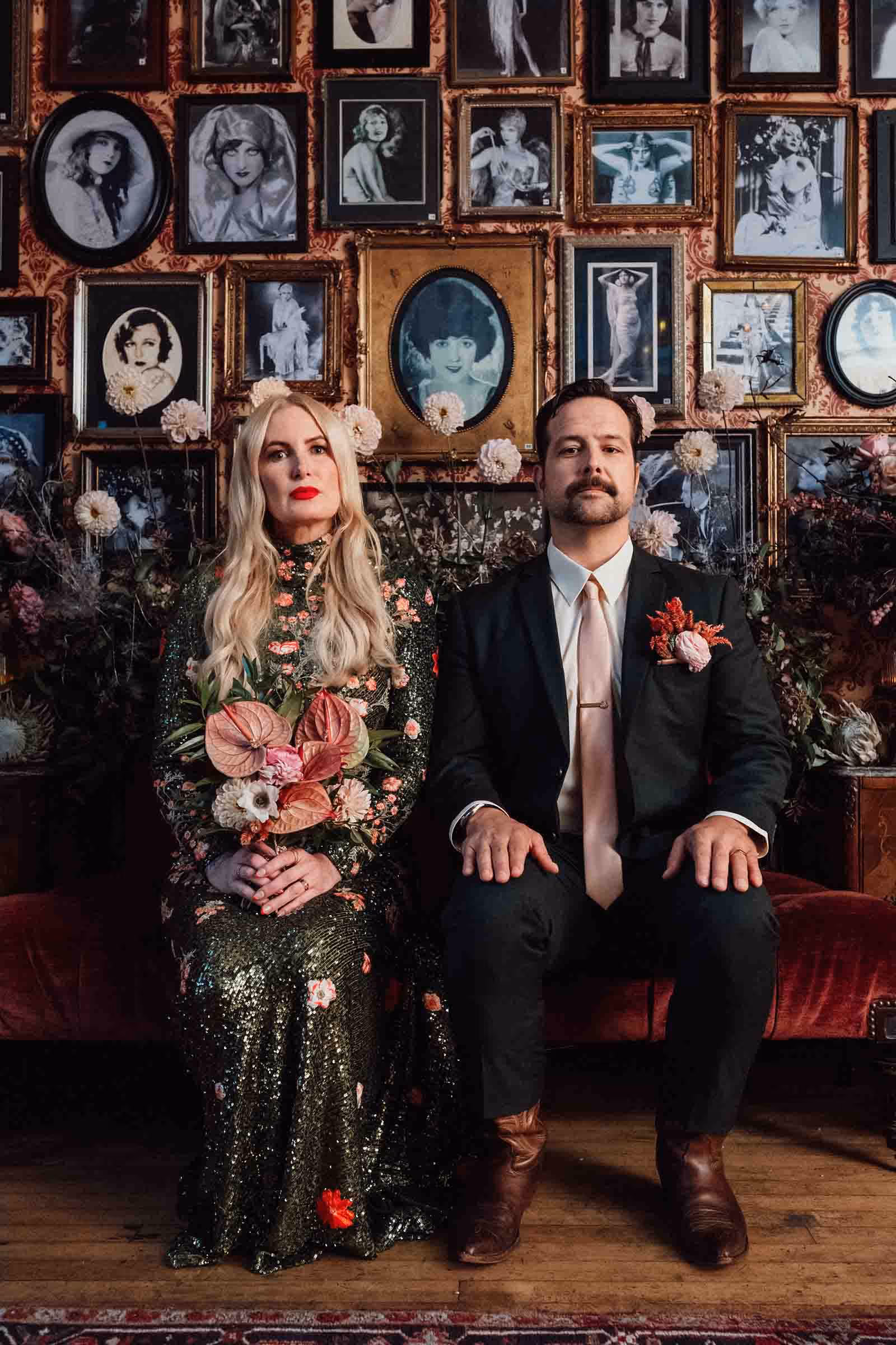 Los Angeles bride and groom sitting side-by-side for an American Gothic inspired portrait on a red velvet couch with vintage photos and frames on the wall behind them.