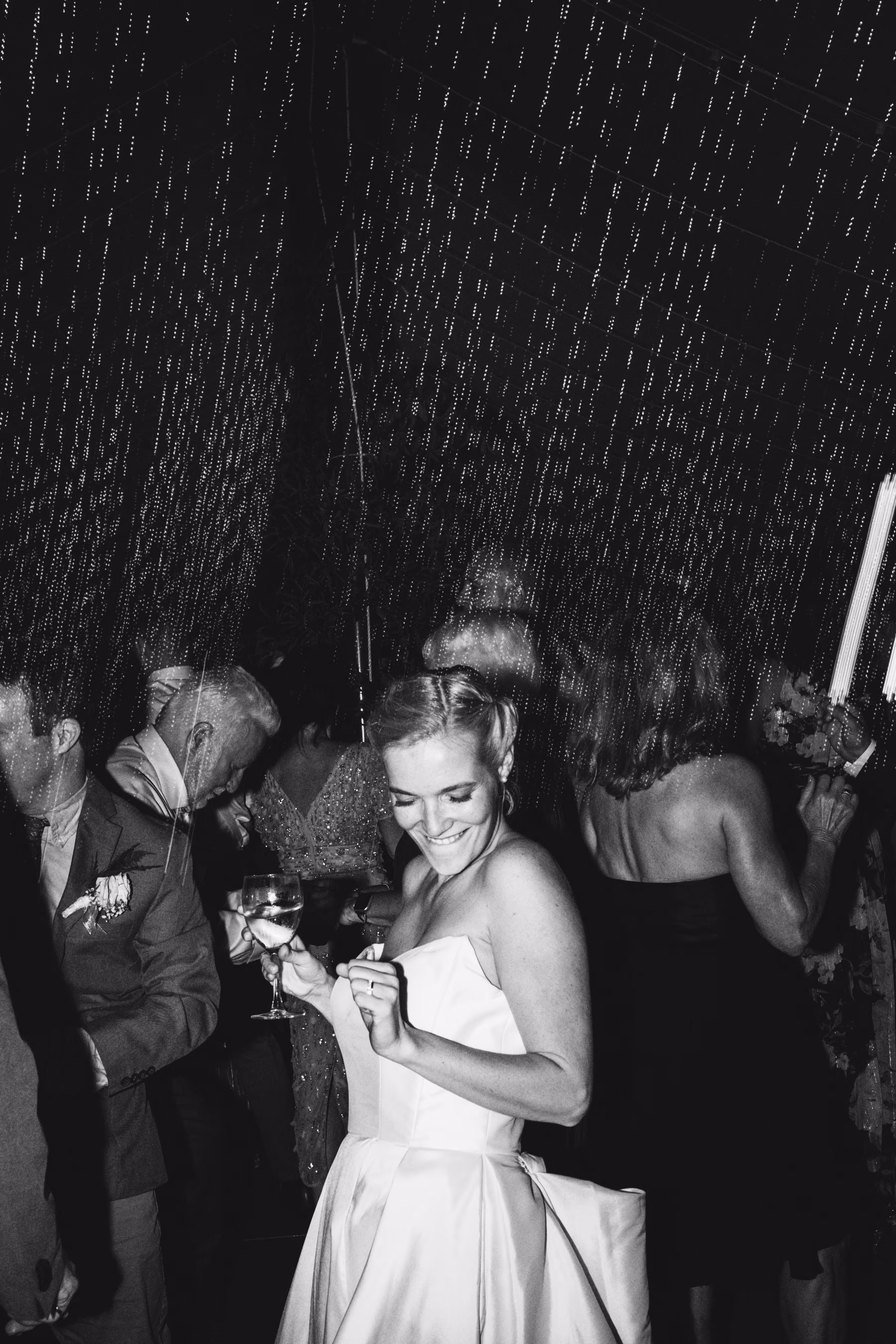 Black and white image of bride looking over shoulder and laughing as friends work on fixing zipper on wedding dress.