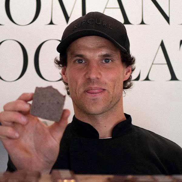 Man wearing a black cap and shirt holding a piece of chocolate close to the camera.