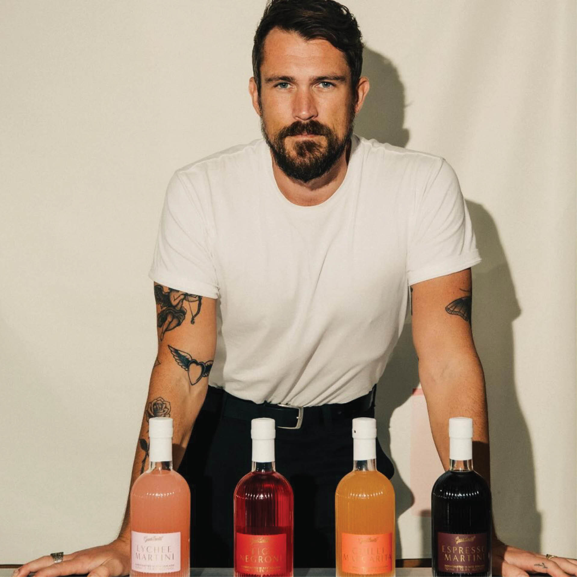 Man with beard and tattoos wearing a white t-shirt standing behind four colorful bottled cocktails on a table.