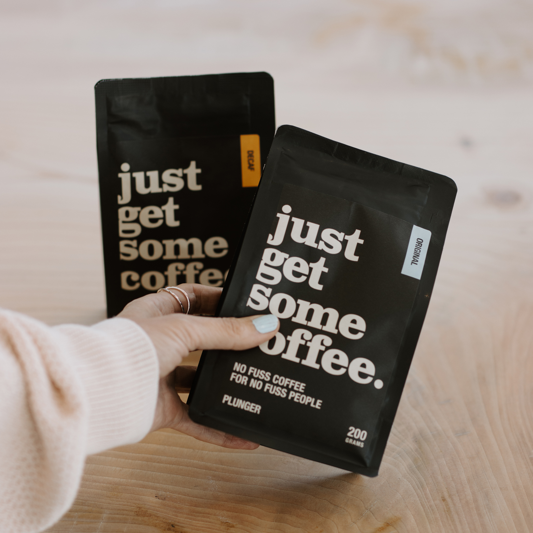 Hand holding black coffee package labeled 'just get some coffee' with text 'No fuss coffee for no fuss people' and 'Plunger 200 grams,' another package labeled decaf in the background.