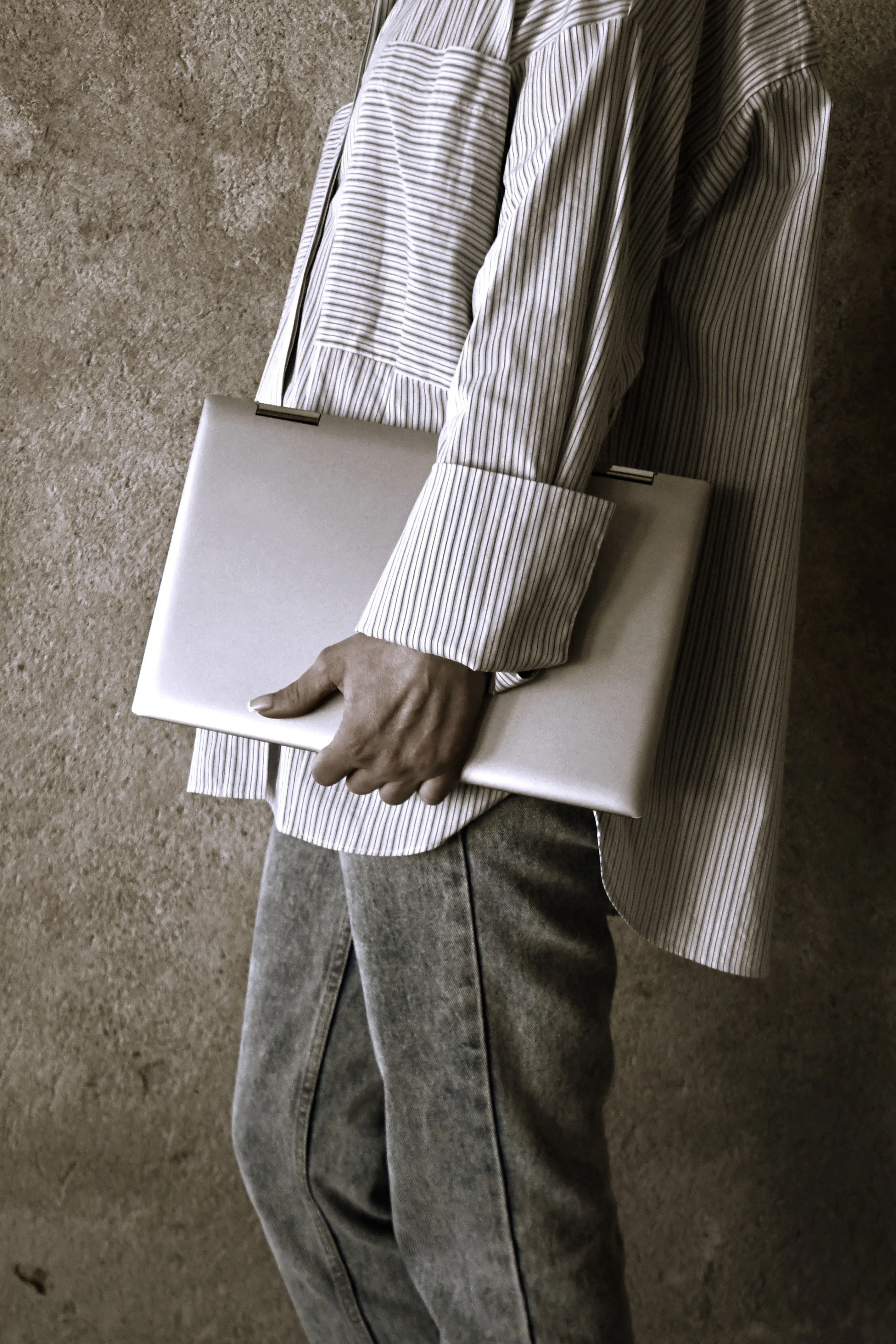 Person tenant un ordinateur portable argenté contre leur corps, portant une chemise à rayures claires et un jean gris délavé.