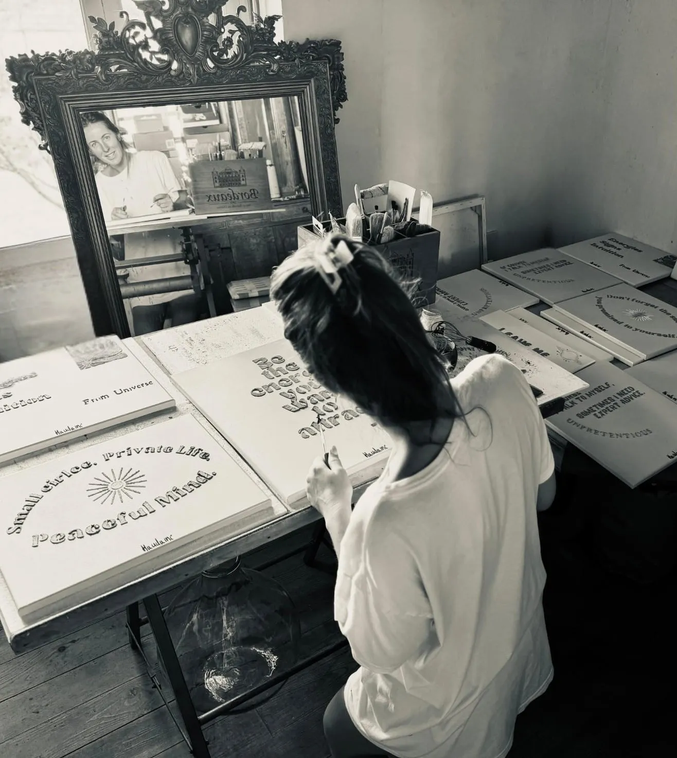 Femme assise à une table, peignant à la main des toiles avec des phrases inspirantes en lettres en relief, reflet dans un grand miroir orné.