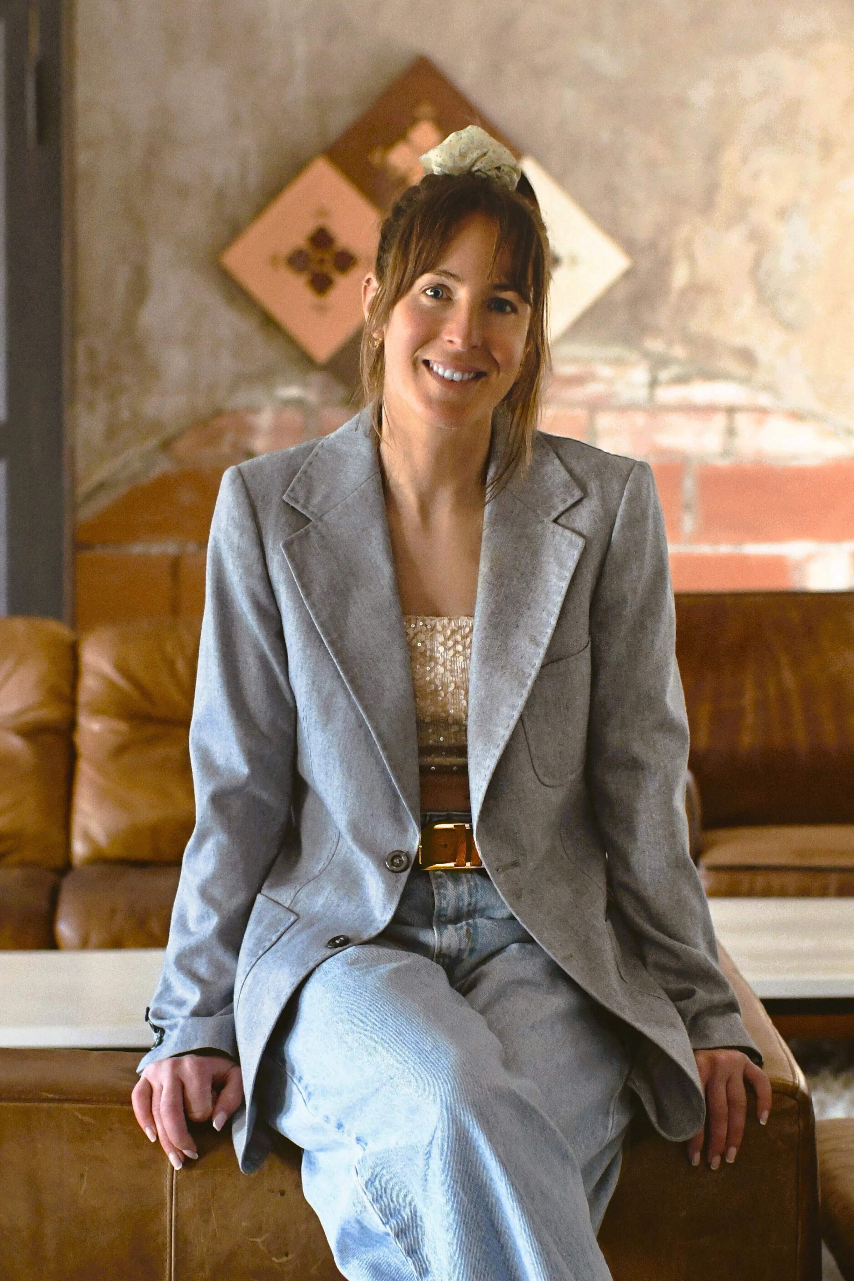 Jeune femme souriante assise sur un canapé en cuir dans un intérieur avec un mur en briques apparentes.