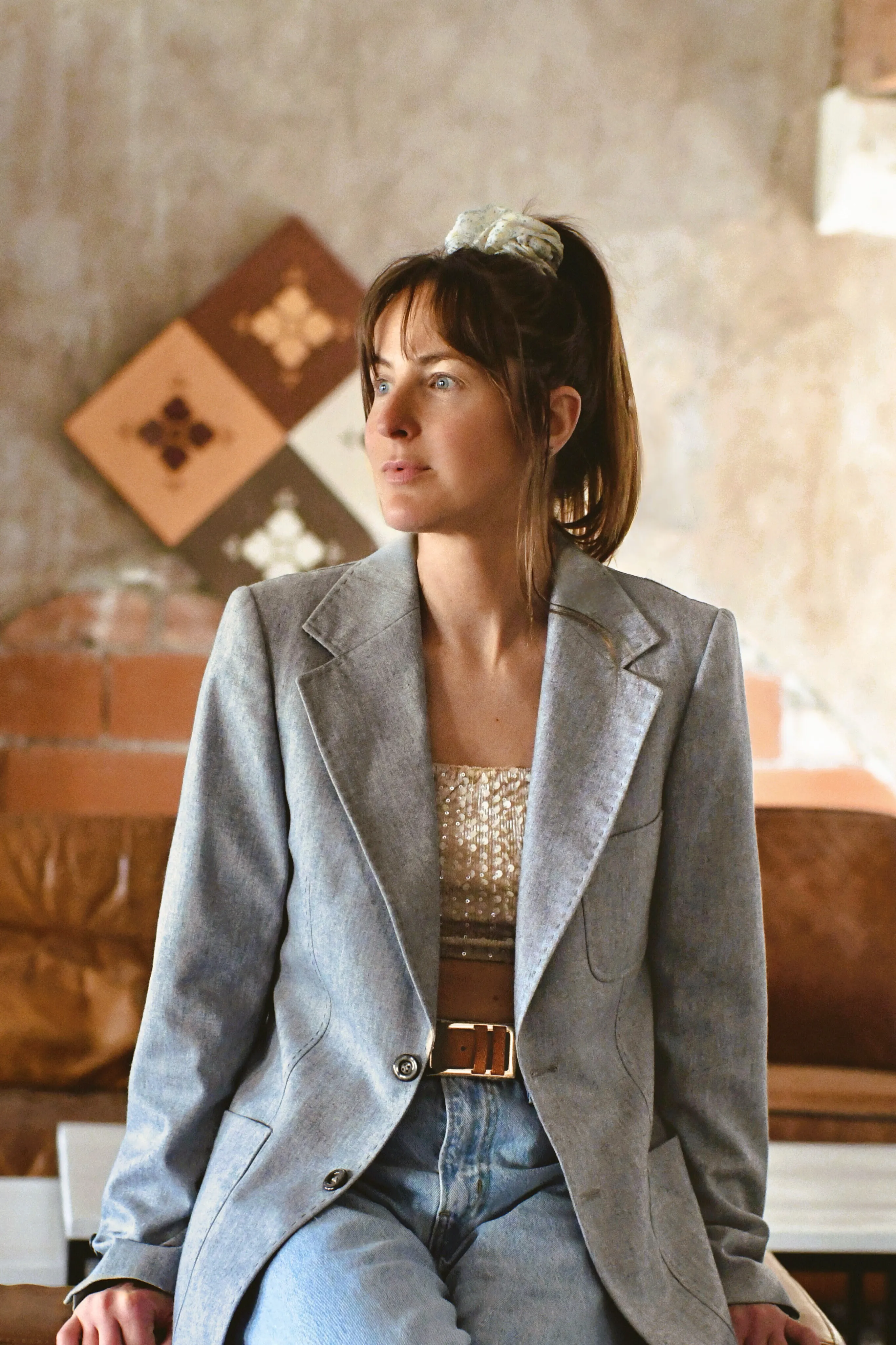 Femme assise portant un blazer gris, un crop top pailleté, et un jean bleu clair, regardant vers la droite.