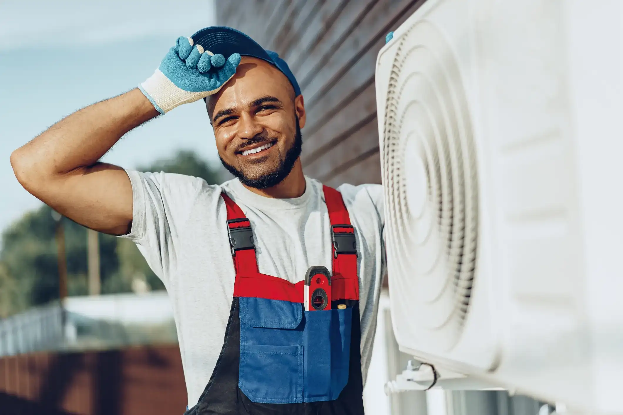 A smiling HVAC technician in overalls and a cap standing next to an outdoor air conditioning unit.