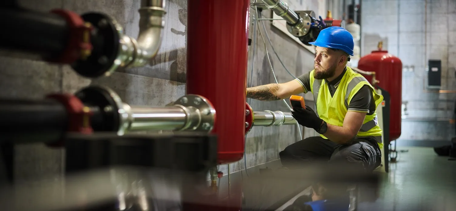 A professional technician wearing a hard hat and high-visibility vest kneels in a large boiler room, holding a meter to check pipes and industrial heating equipment.