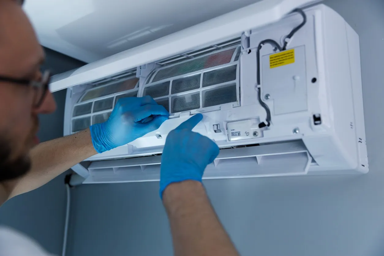 A technician wearing blue gloves performs maintenance on the internal components of an open wall-mounted ductless mini-split unit. The unit's front cover is raised, revealing the internal mesh filters and wiring.