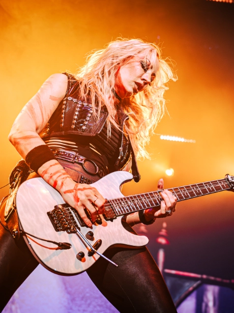 Guitariste féminine avec cheveux blonds longs jouant une guitare électrique blanche sur scène, illuminée par une lumière orange intense.