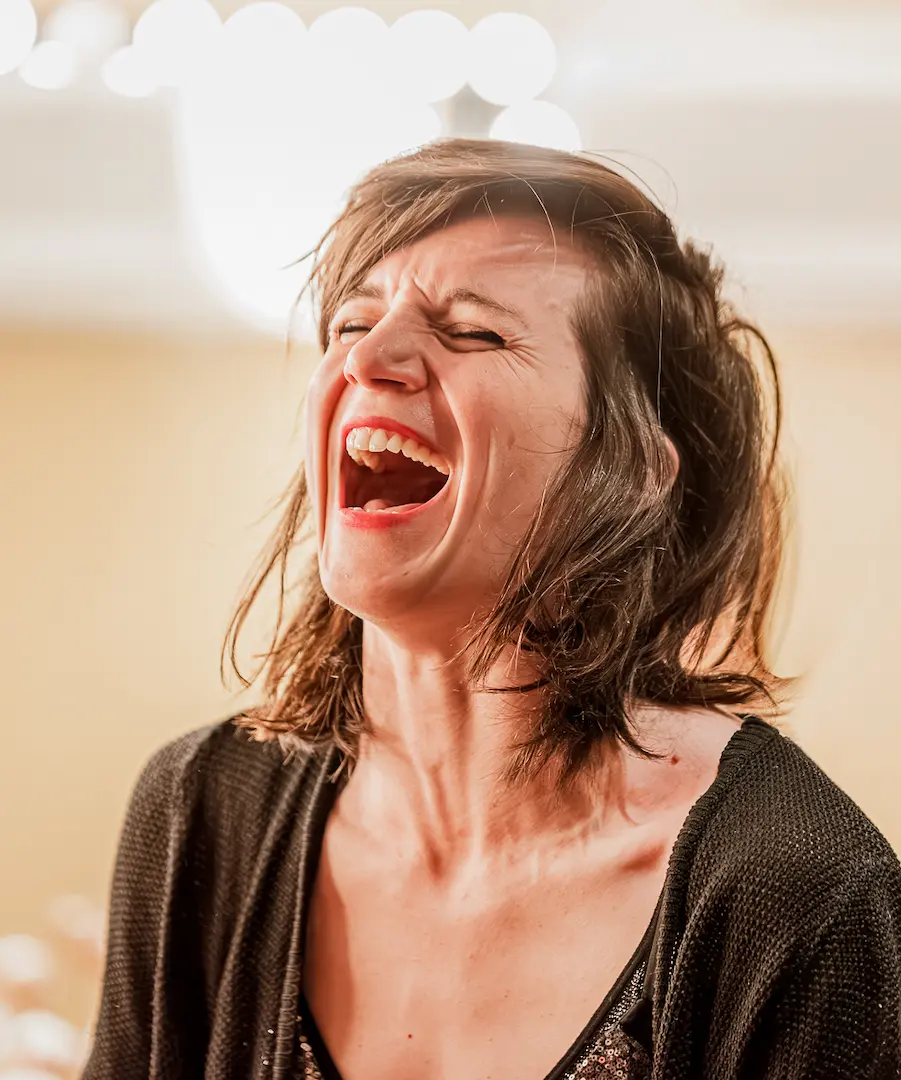 Femme aux cheveux bruns riant aux éclats avec un pull noir et un haut à paillettes. Coulisses Les Franglaises