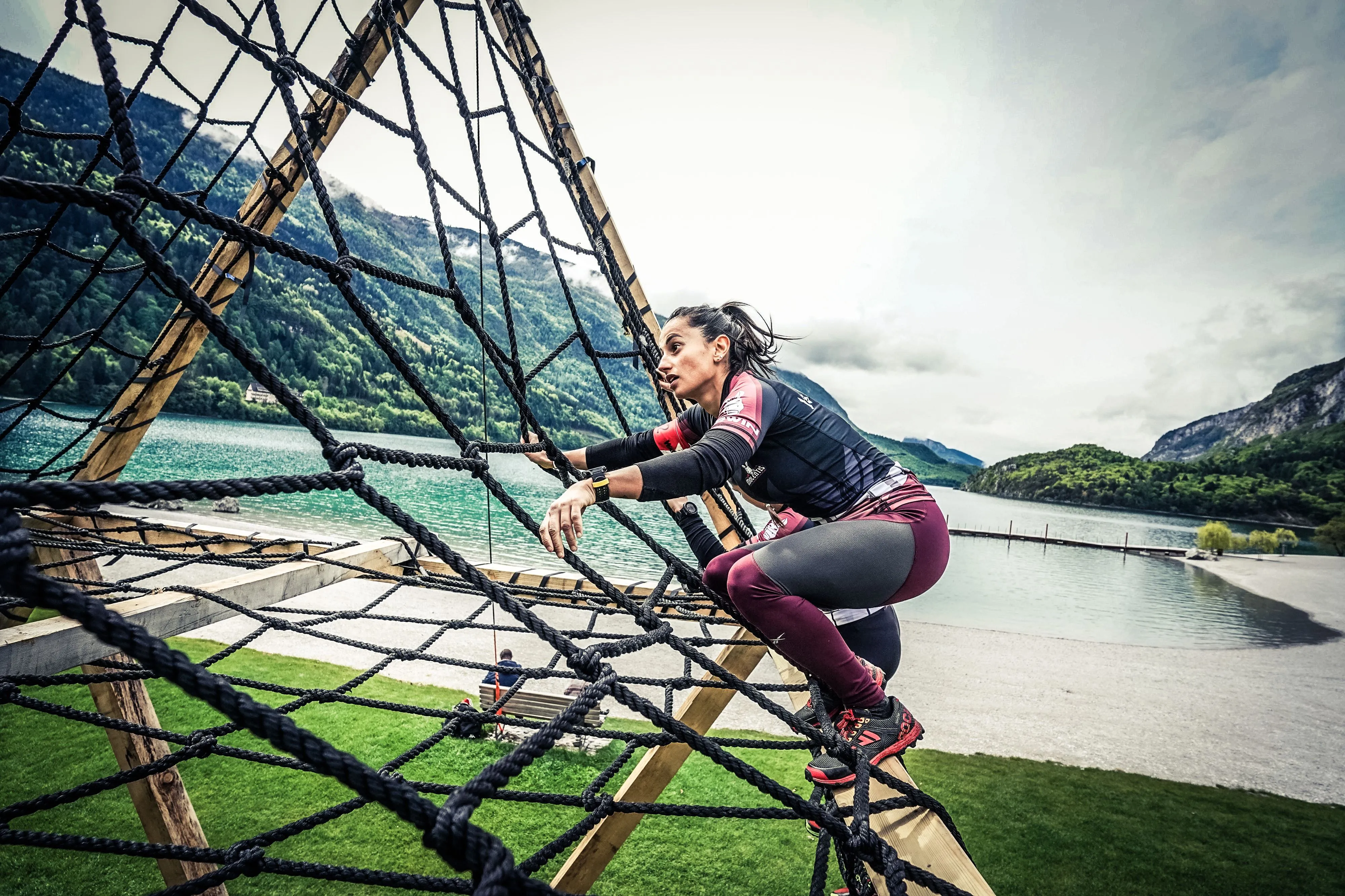 Female athlete climbing the cargo net pyramid beside Lake Molveno at the X-Warriors OCR race