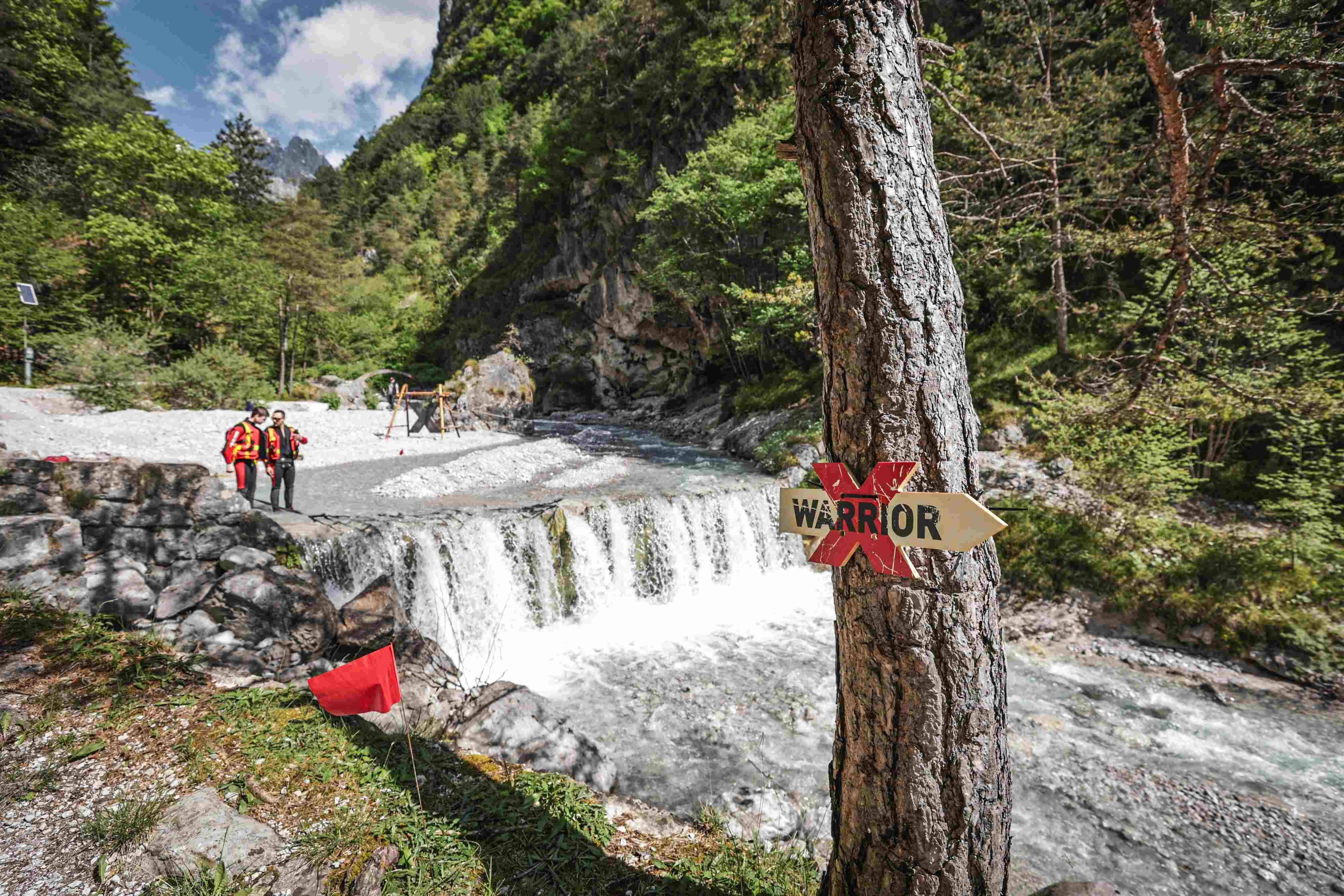 Two men climbing a log in the water with many rocks and stones in the background during the X Warriors race.
