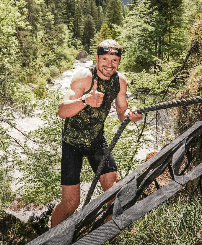Smiling athlete giving a thumbs up on the rope climb above the river at X-Warriors Molveno