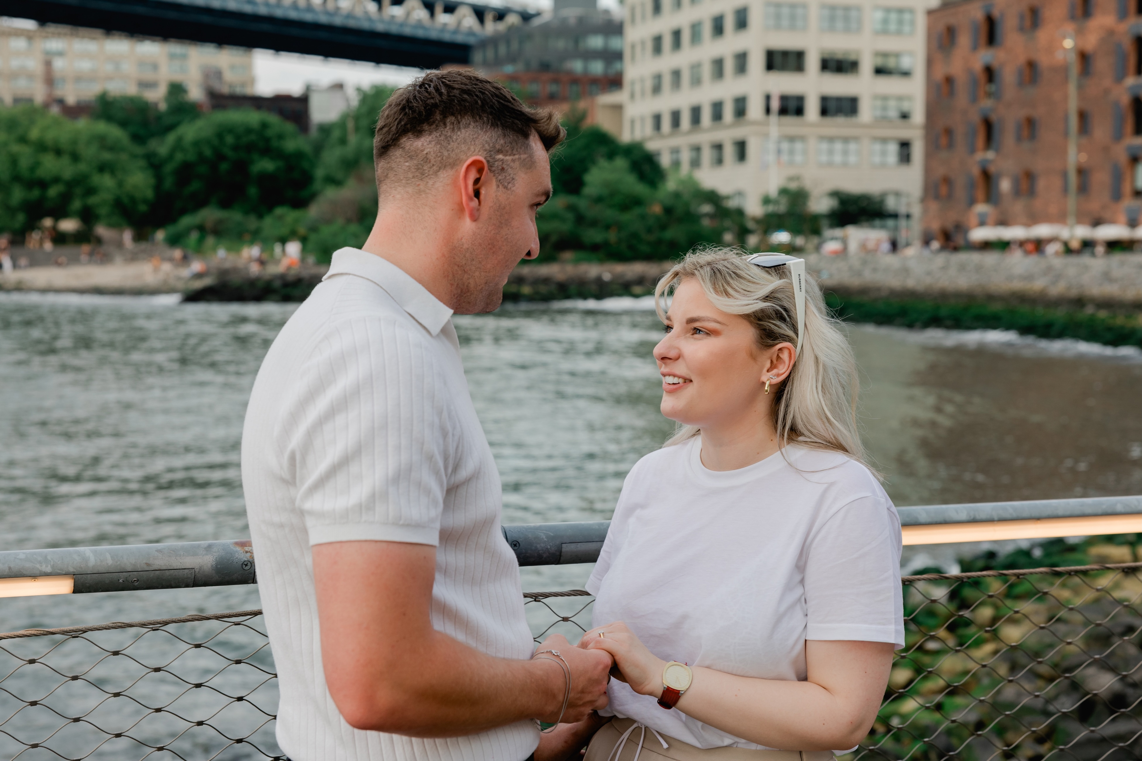 Couple holding hands on the waterfront at Brooklyn Bridge Park after surprise proposal. 