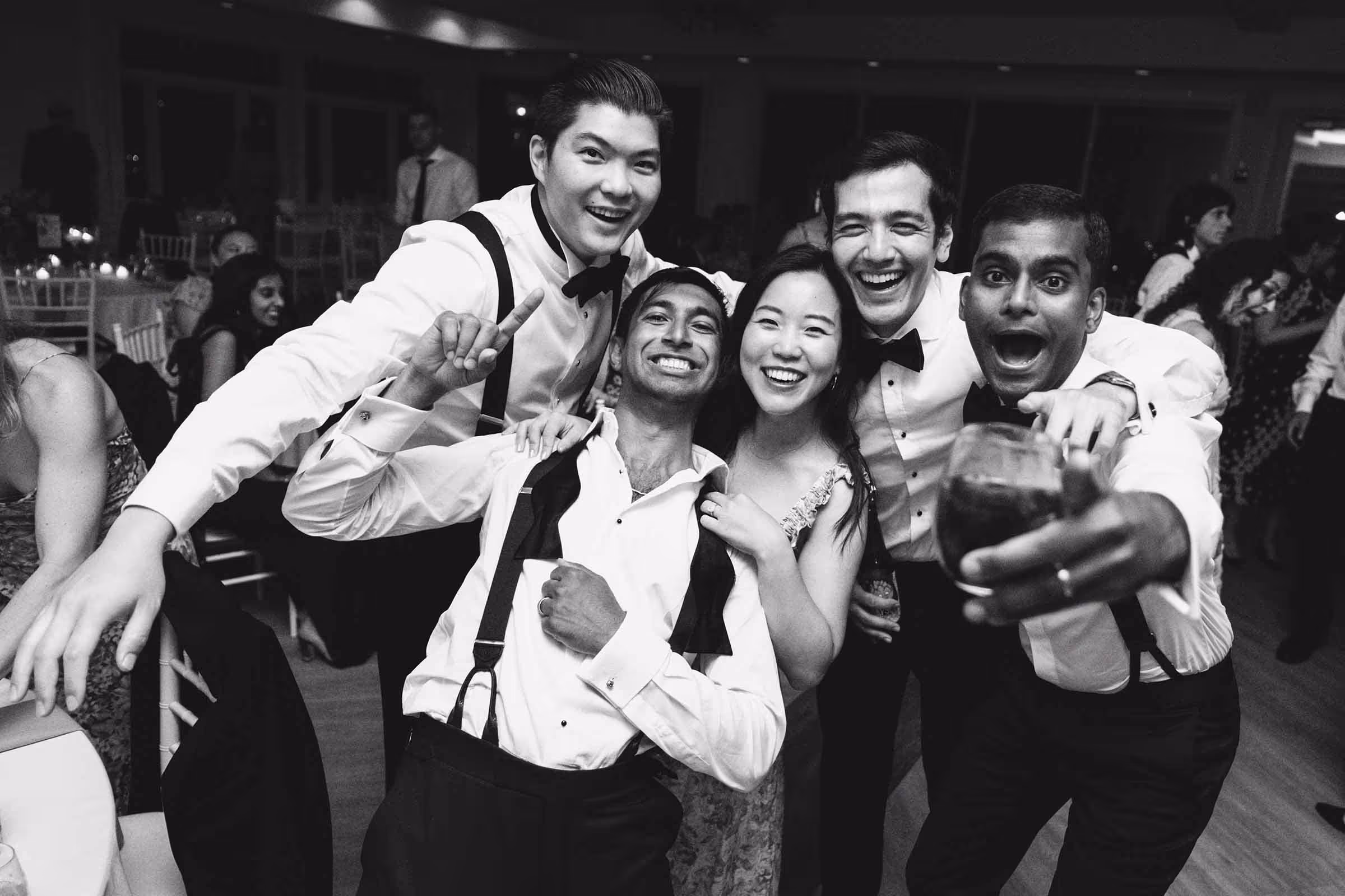 Groom and friends party on the dance floor while interacting with the camera, smiling and pointing.
