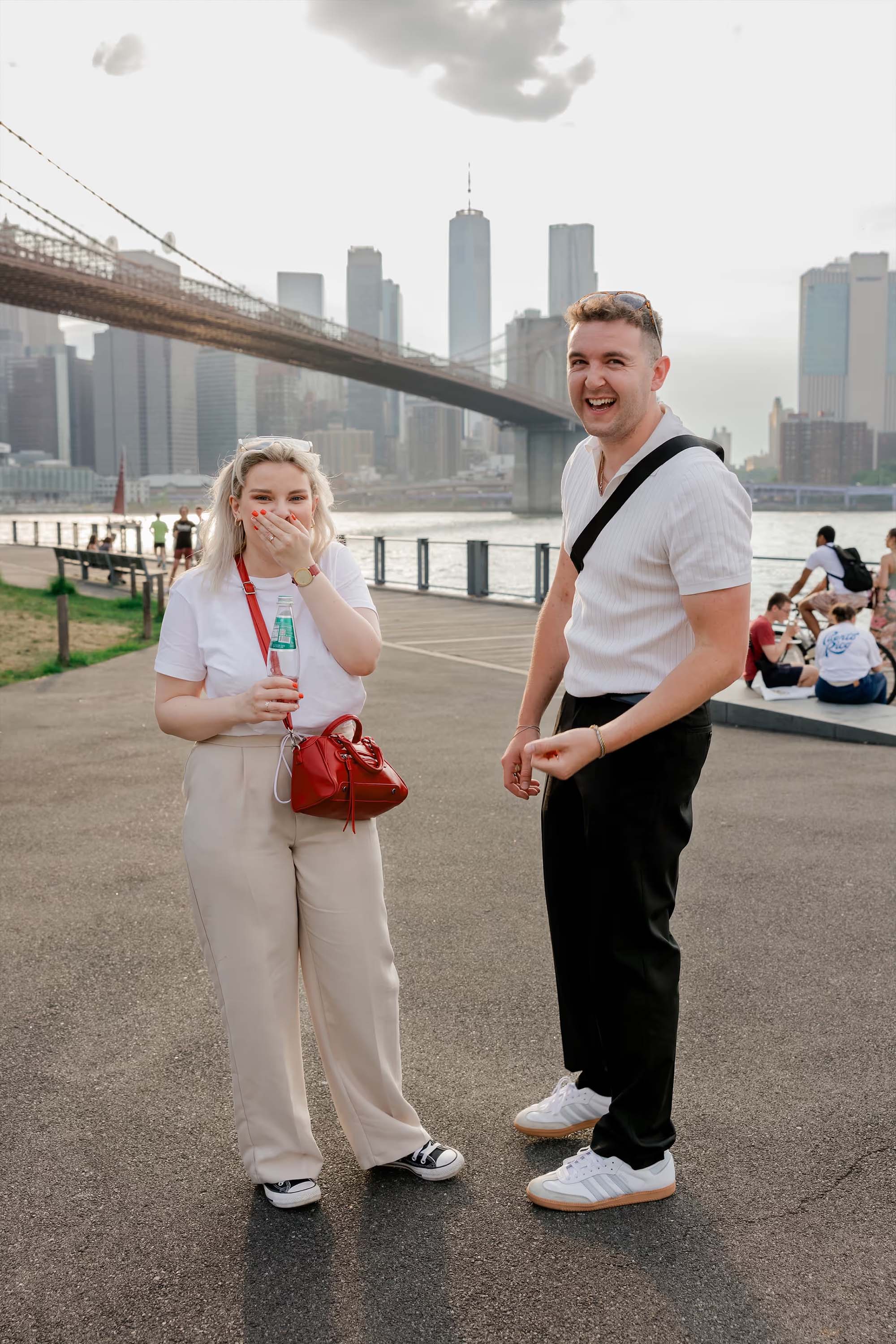 Couple reacts to the camera after a surprise proposal in DUMBO's Brooklyn Bridge Park with the Manhattan bridge and city skyline behind them 