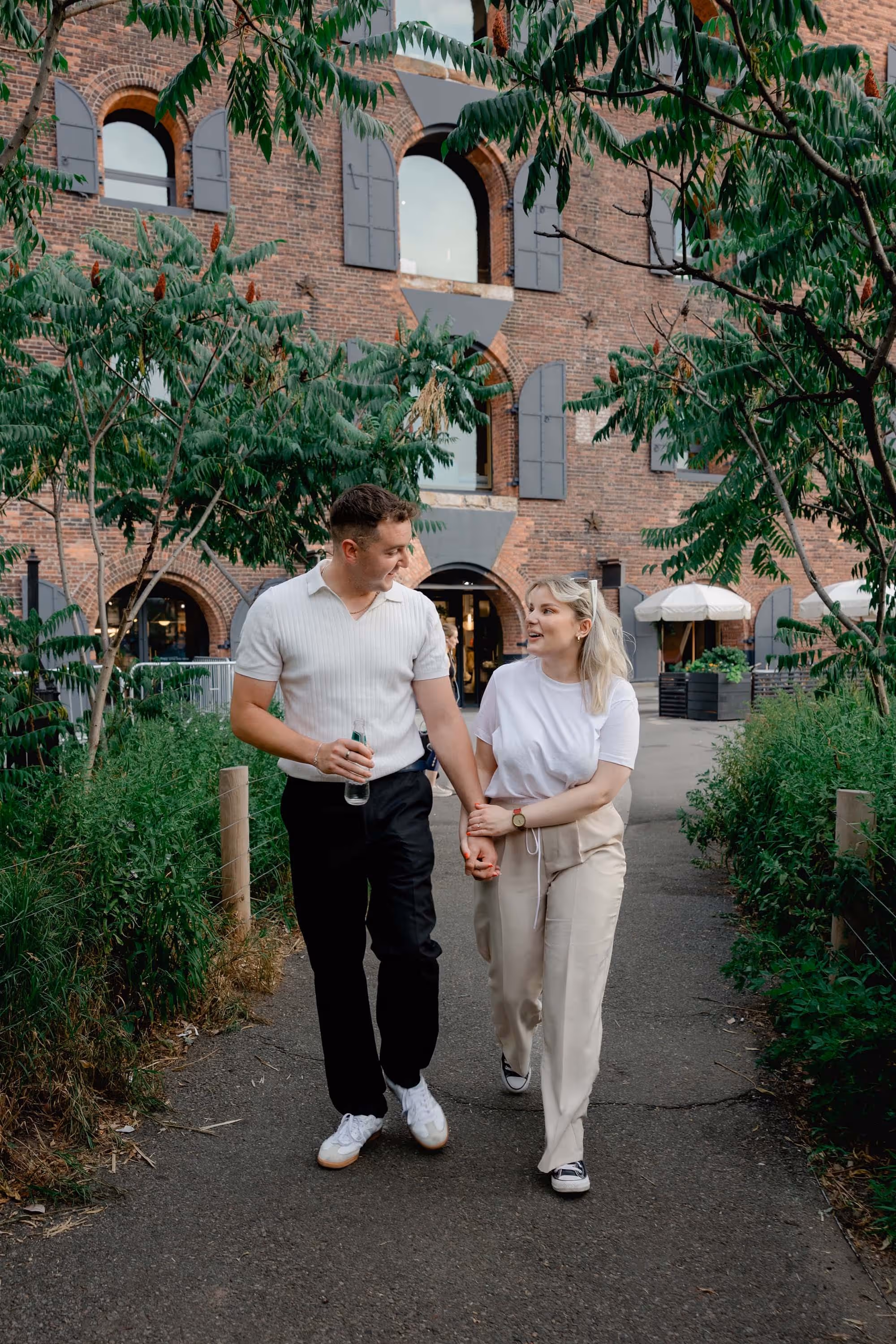 Couple holds hands while walking through the greenery at Brooklyn Bridge Park in DUMBO following a surprise marriage proposa    