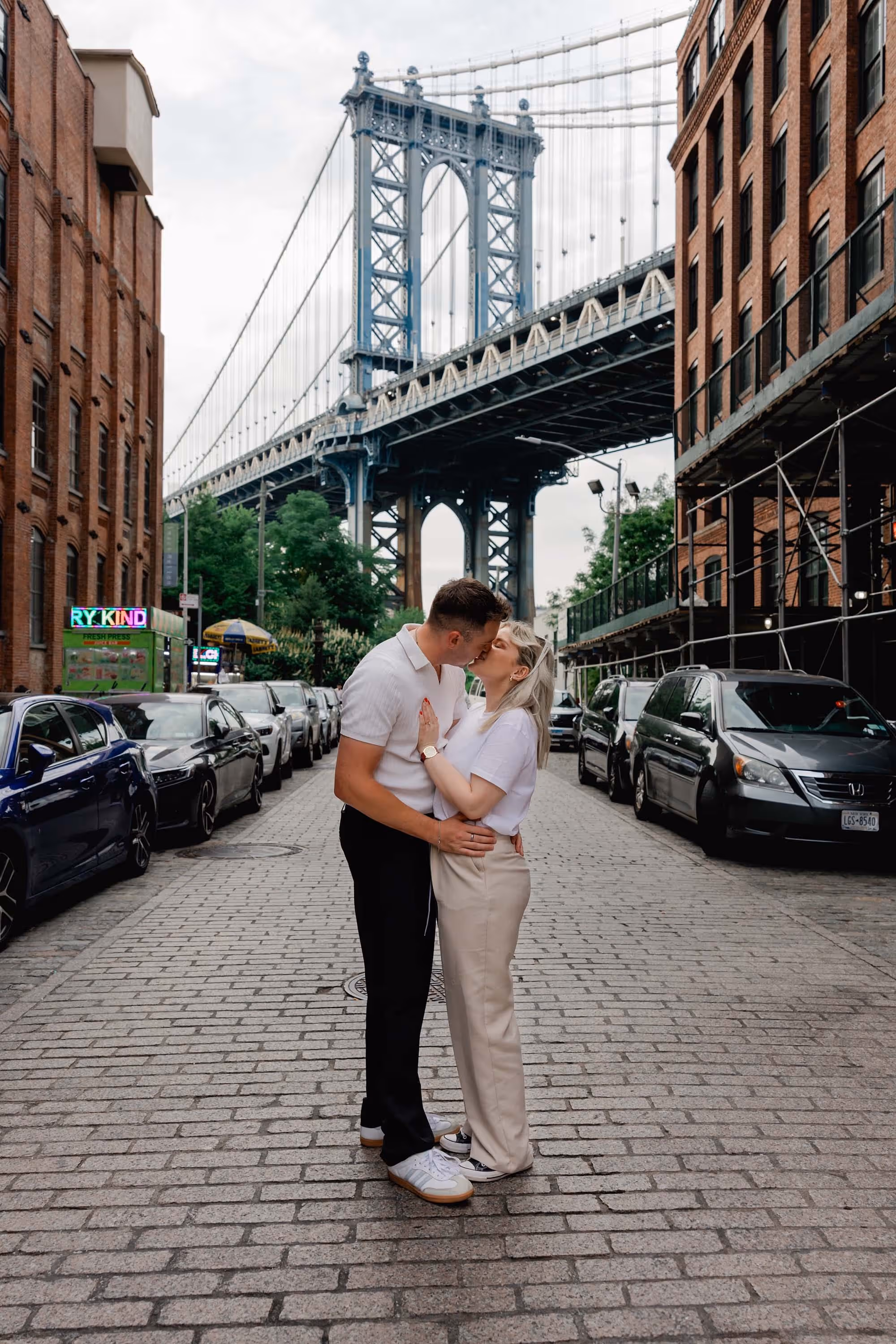 Couple from the UK kiss on the cobblestone streets of DUMBO after a surprise proposal with the Brooklyn Bridge behind them