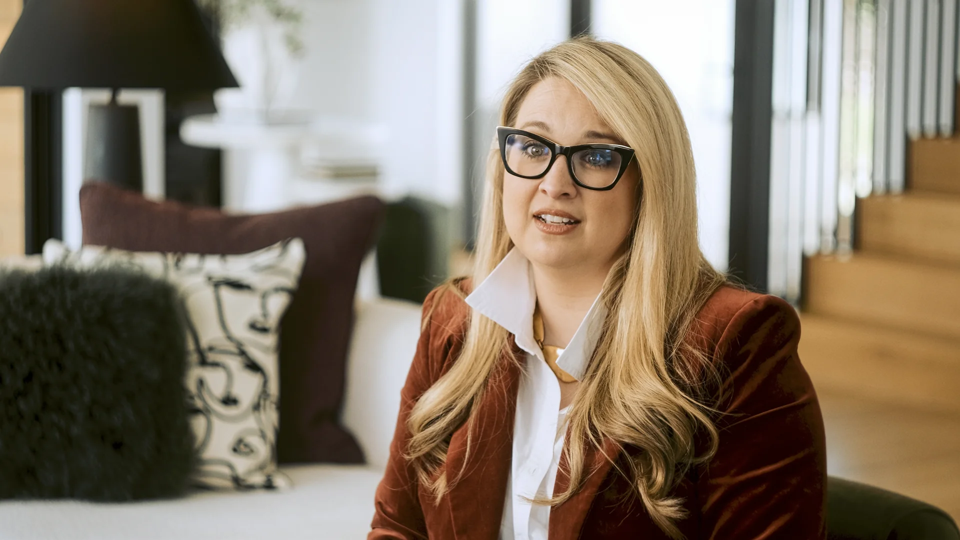 Blonde woman with glasses and long hair wearing a brown jacket and white shirt sitting on a sofa indoors.