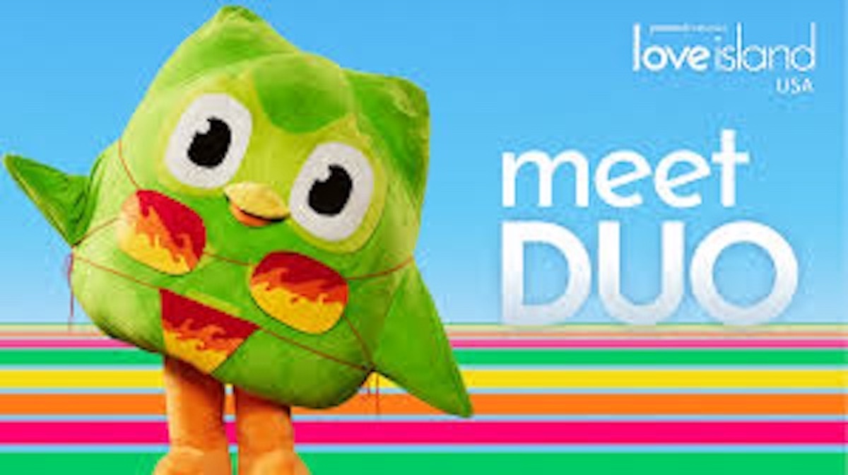 A large, green owl mascot with big eyes and orange feet stands on a colorful striped background next to the text love island USA and meet DUO.