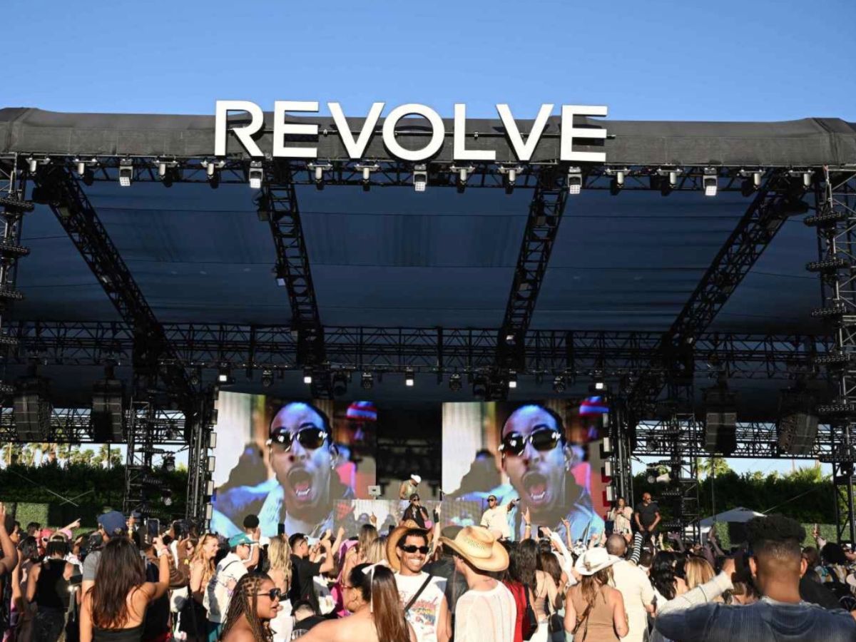 A large outdoor music stage with REVOLVE on top, big screens displaying a performer, and a crowd of people enjoying the event under a clear blue sky.