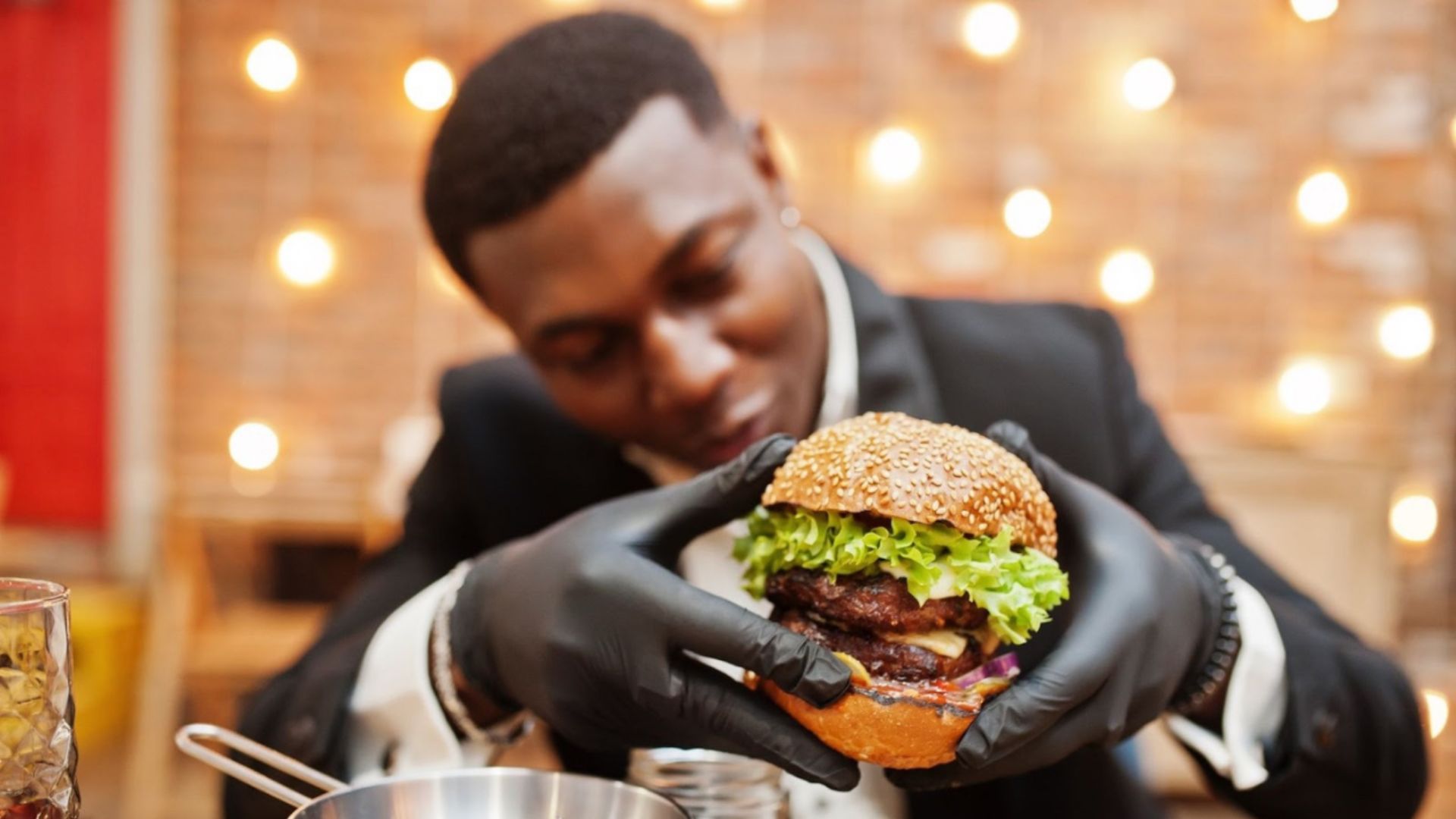 A person wearing a suit and black gloves holds a large sesame seed burger filled with lettuce, tomato, and a beef patty, with a blurred background of warm string lights.