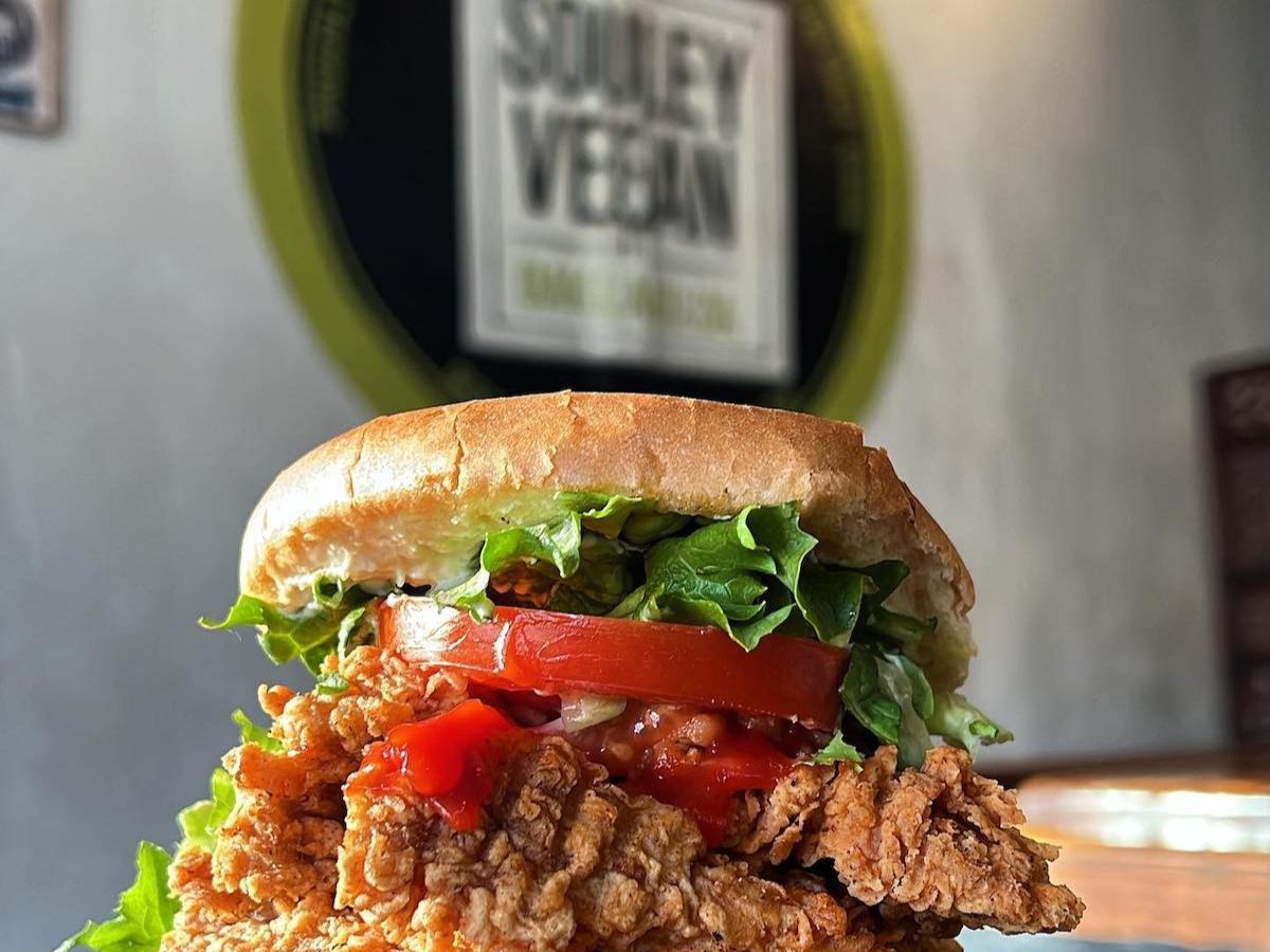 A close-up of a crispy vegan fried chicken sandwich with lettuce, tomato, onions, and sauce, served on a bun. A blurred “Souley Vegan” sign is visible in the background.A close-up of a crispy vegan fried chicken sandwich with lettuce, tomato, onions, and sauce, served on a bun. A blurred “Souley Vegan” sign is visible in the background.