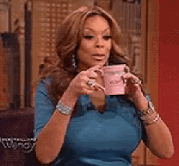 A woman with long hair and a blue top holds a pink cup with both hands, raising her eyebrows and looking to the side with a surprised or dramatic expression.