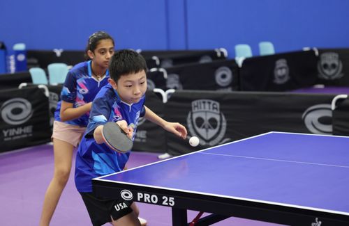 Two young table tennis players in blue uniforms, one about to hit the ball with a paddle in an indoor tournament setting.
