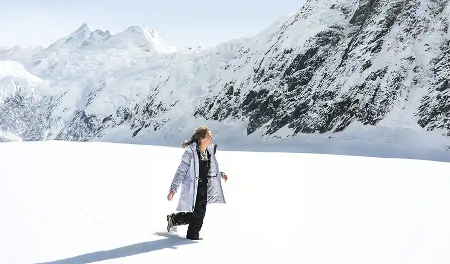 A person walks across a vast snowy landscape, wearing a silver parka and boots, surrounded by towering, snow-covered mountains under bright sunlight.
