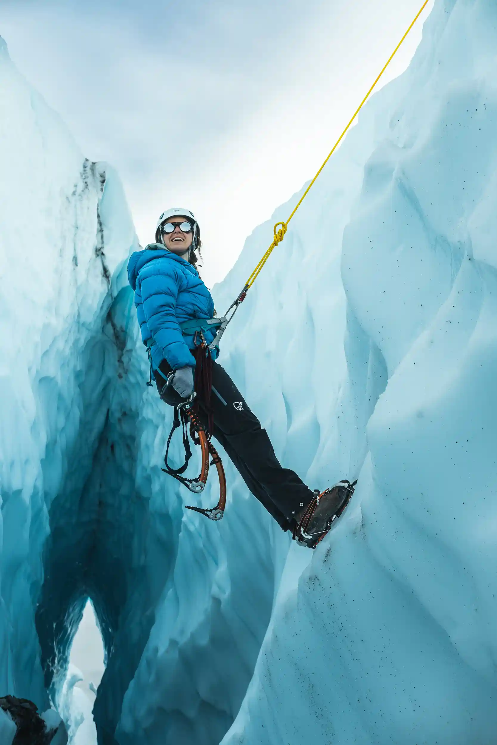 A smiling ice climber wearing a blue puffer jacket, helmet, and sunglasses ascends a steep wall of blue glacial ice using crampons and ice axes. A bright yellow rope secures them as they lean back mid-climb, surrounded by towering frozen walls and a deep crevasse below.