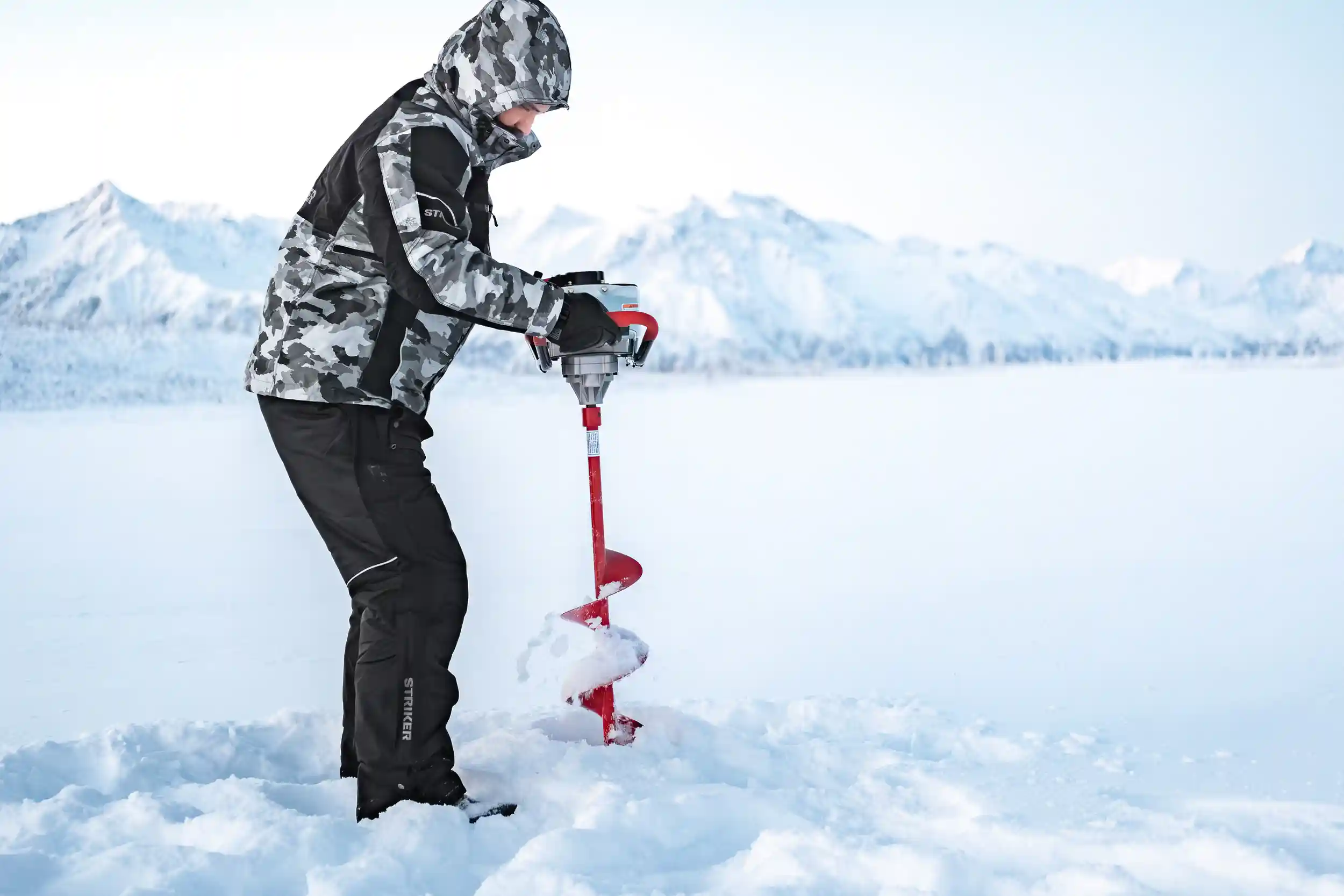 A person wearing a gray camo jacket and black snow pants labeled “Striker” operates a red ice auger on a frozen lake surrounded by snow-covered mountains. The scene captures the crisp, bright atmosphere of a winter day in Alaska, with fine snow spraying as the auger cuts into the ice.