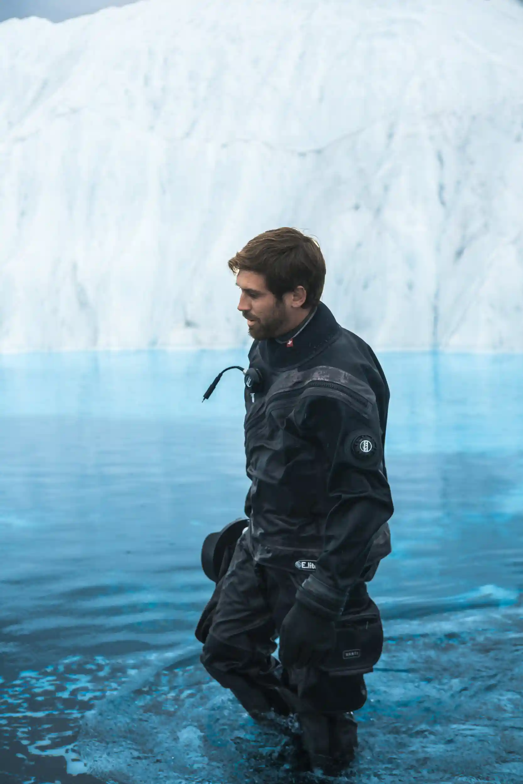 A man in a black drysuit wades through icy blue glacial water, holding his underwater camera in one hand. Behind him rises a massive wall of white and blue ice, reflecting softly on the still surface, creating a powerful image of exploration and endurance in Alaska’s frozen landscape.