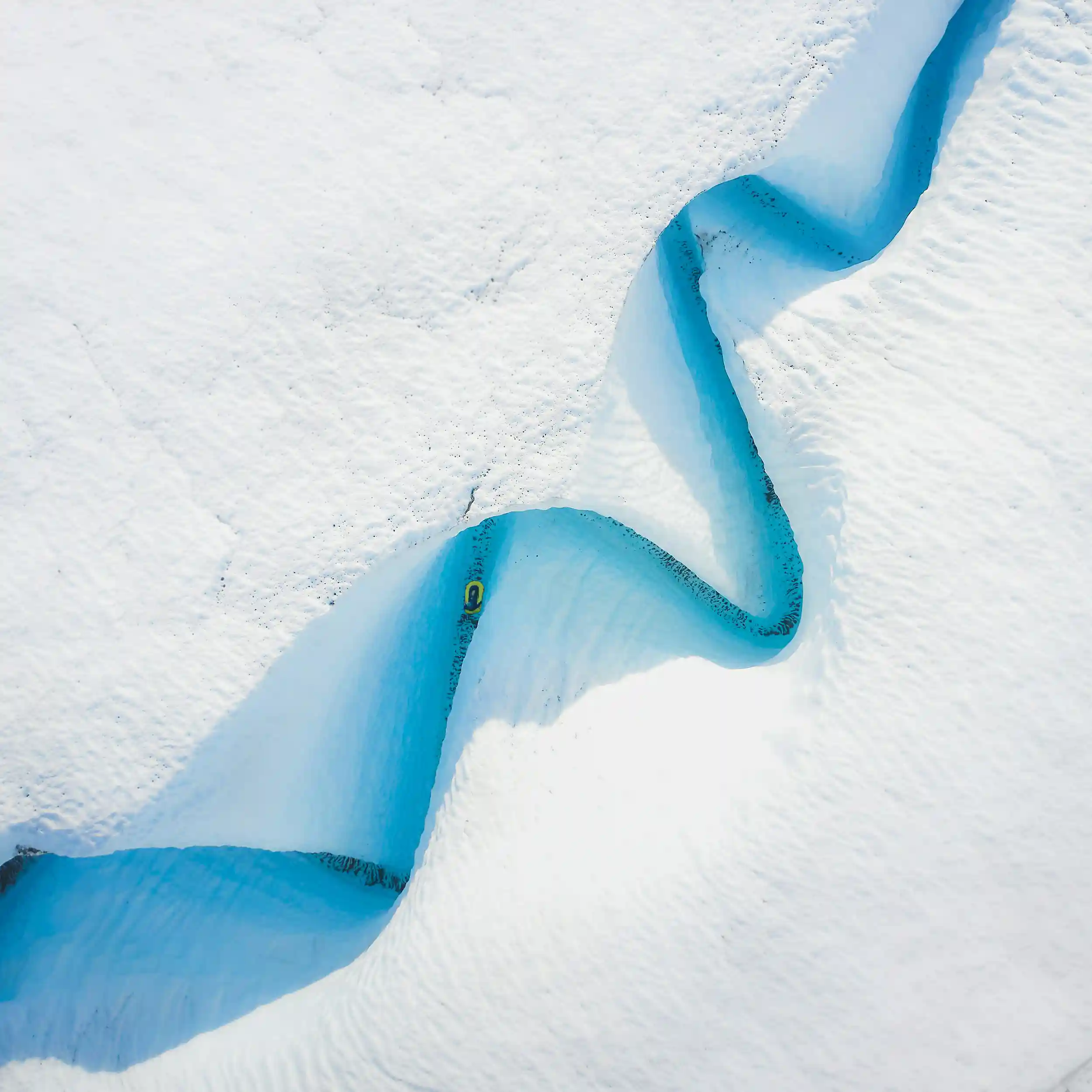 A tiny yellow packraft floats through a winding, deep-blue melt channel carved into a vast white glacier. Viewed from above, the river-like path snakes across the ice, creating a striking contrast between the brilliant blue water and the bright, textured snow surrounding it.