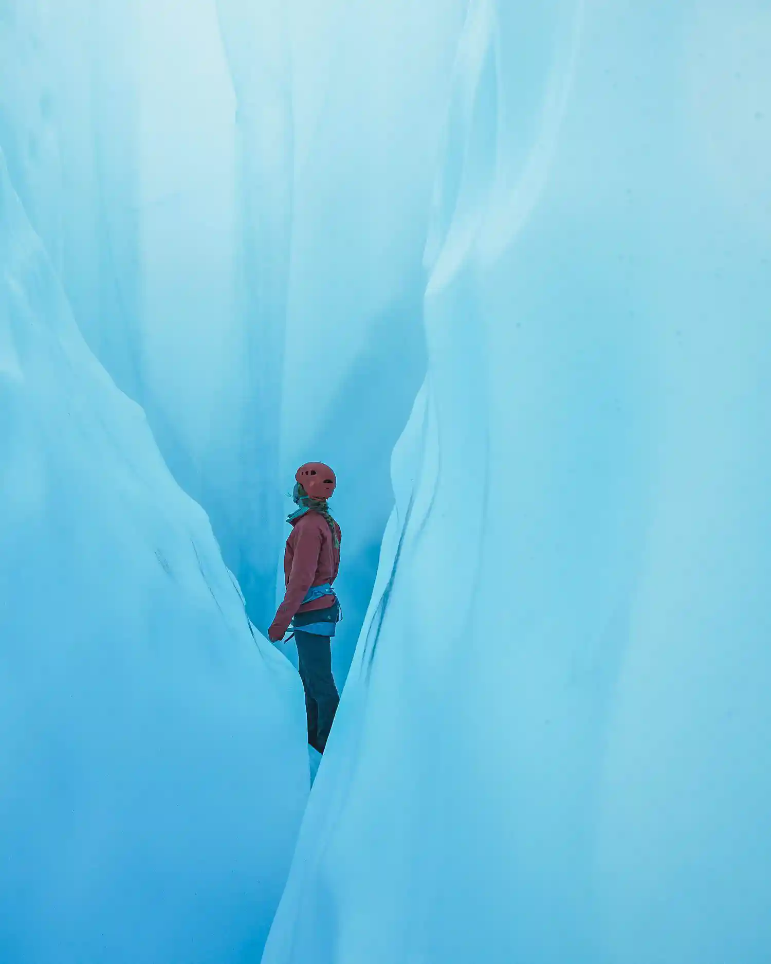 A climber wearing a red jacket, helmet, and harness stands deep within a narrow blue glacial crevasse, surrounded by smooth, towering ice walls glowing with soft light. The scene captures the scale and beauty of Alaska’s frozen landscape and the sense of awe in the moment.