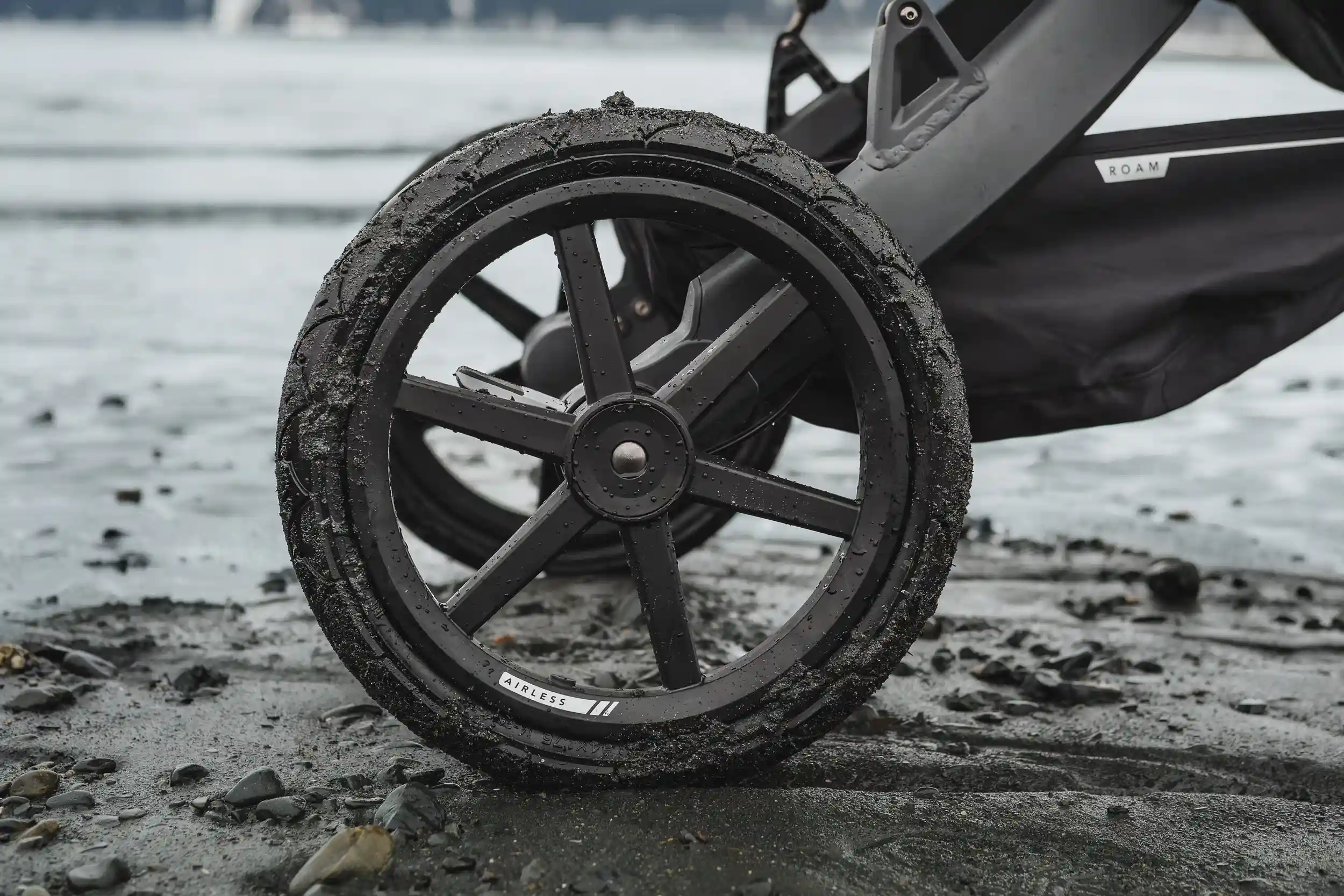 A detailed close-up of a black Guava stroller wheel coated with sand and water droplets, resting on wet pebbled beach terrain near the ocean. The tire label reads “AIRLESS,” emphasizing its rugged, all-terrain design built for outdoor adventure.
