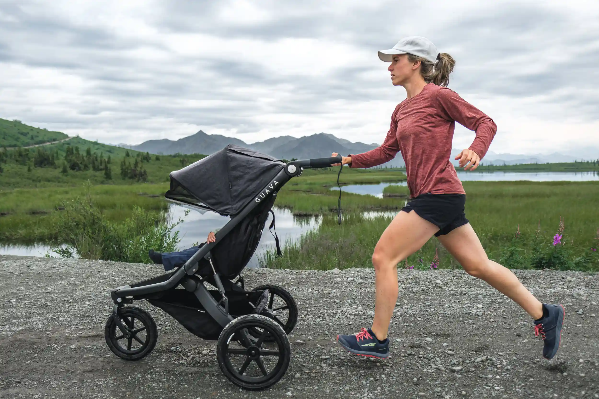 A woman jogs along a gravel trail pushing a black Guava stroller beside a reflective pond surrounded by lush green tundra and distant mountain peaks. She wears a red long-sleeve shirt, black shorts, running shoes, and a light gray cap, captured mid-stride in an active, determined motion under overcast skies.