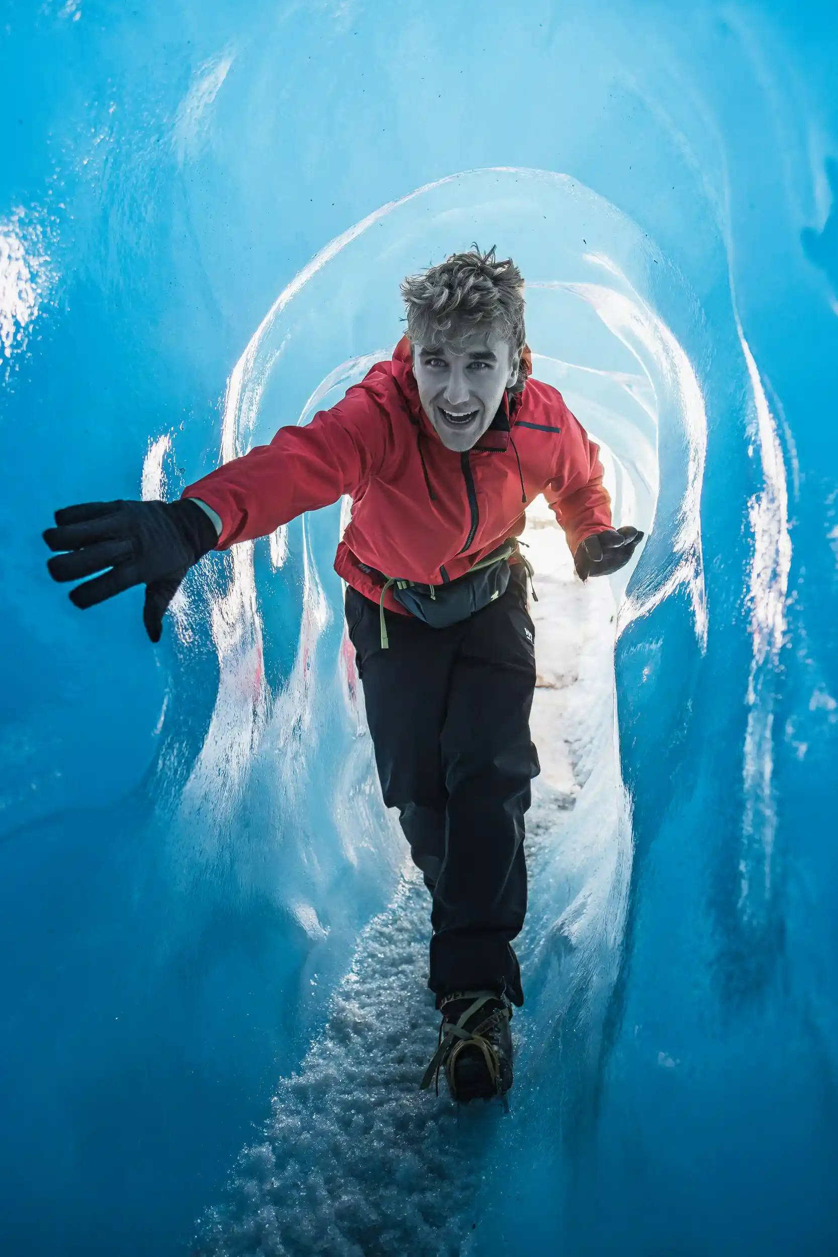 Man in a red jacket walking through a blue ice cave on Mendenhall Glacier