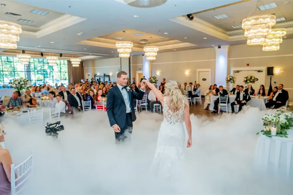 Bride and groom dancing on a cloudlike fog floor with guests watching in a chandelier-lit wedding reception hall.