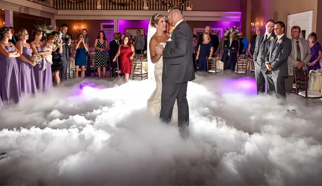 Bride and groom dancing on a cloud-like fog floor surrounded by wedding guests in a decorated venue.