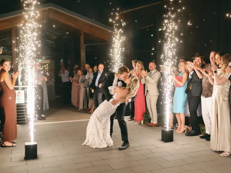 Bride and groom share a kiss while guests clap and cold spark fountains light up the night at an outdoor wedding.
