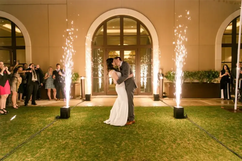 Bride and groom kissing outdoors on grass with wedding guests watching and cold spark fountains sparkling around them.
