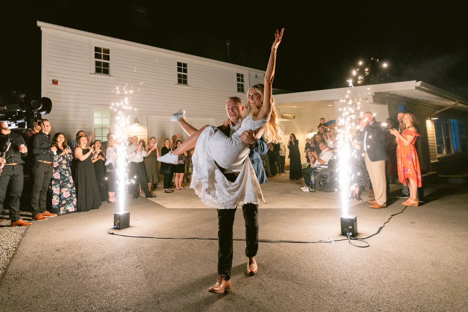 Groom carrying bride in a white dress celebrating at night outdoors with guests clapping and sparklers lighting the scene.