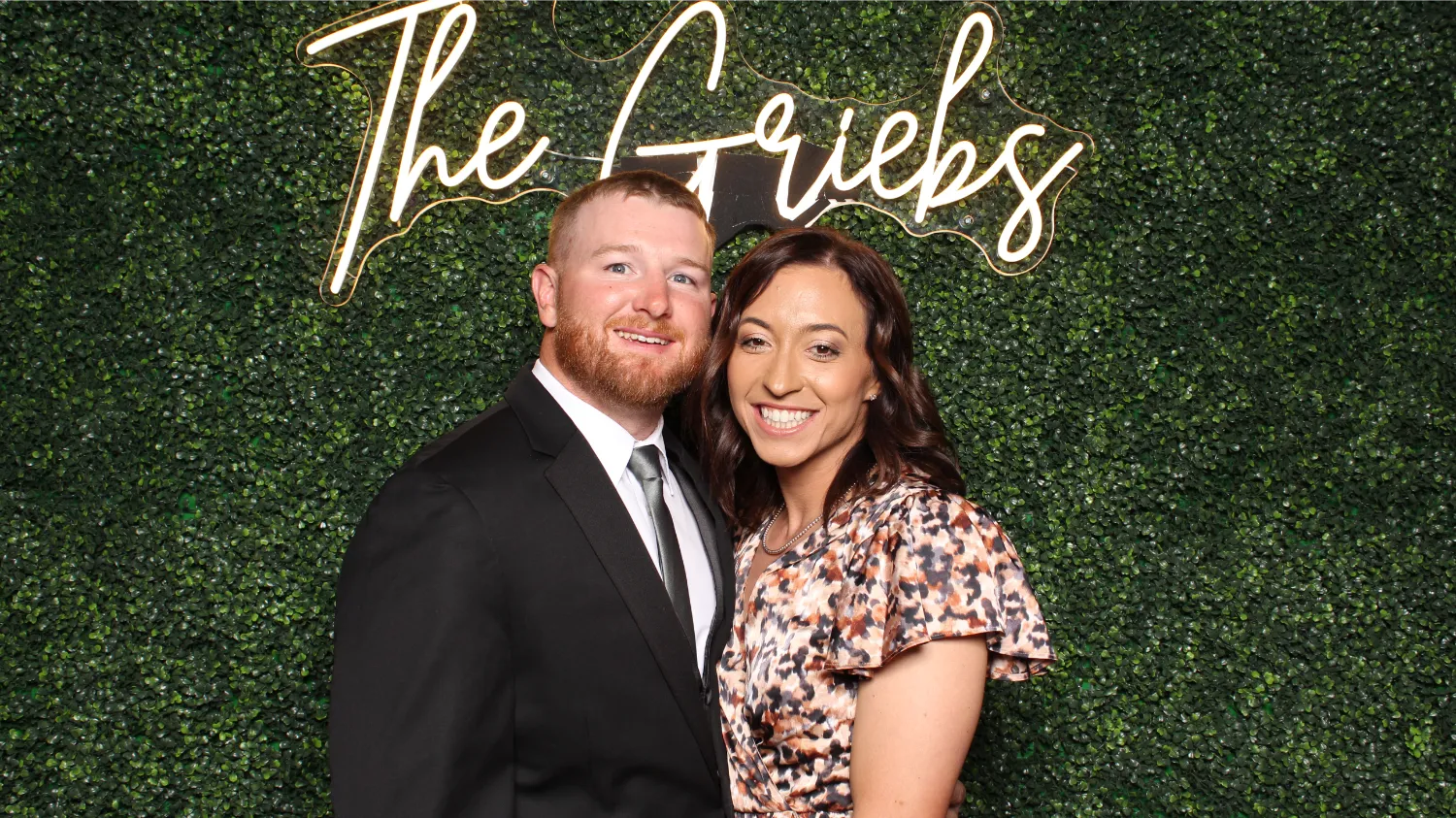 Smiling couple dressed formally posing in front of a green leafy backdrop with illuminated cursive text saying 'The Griebs'.