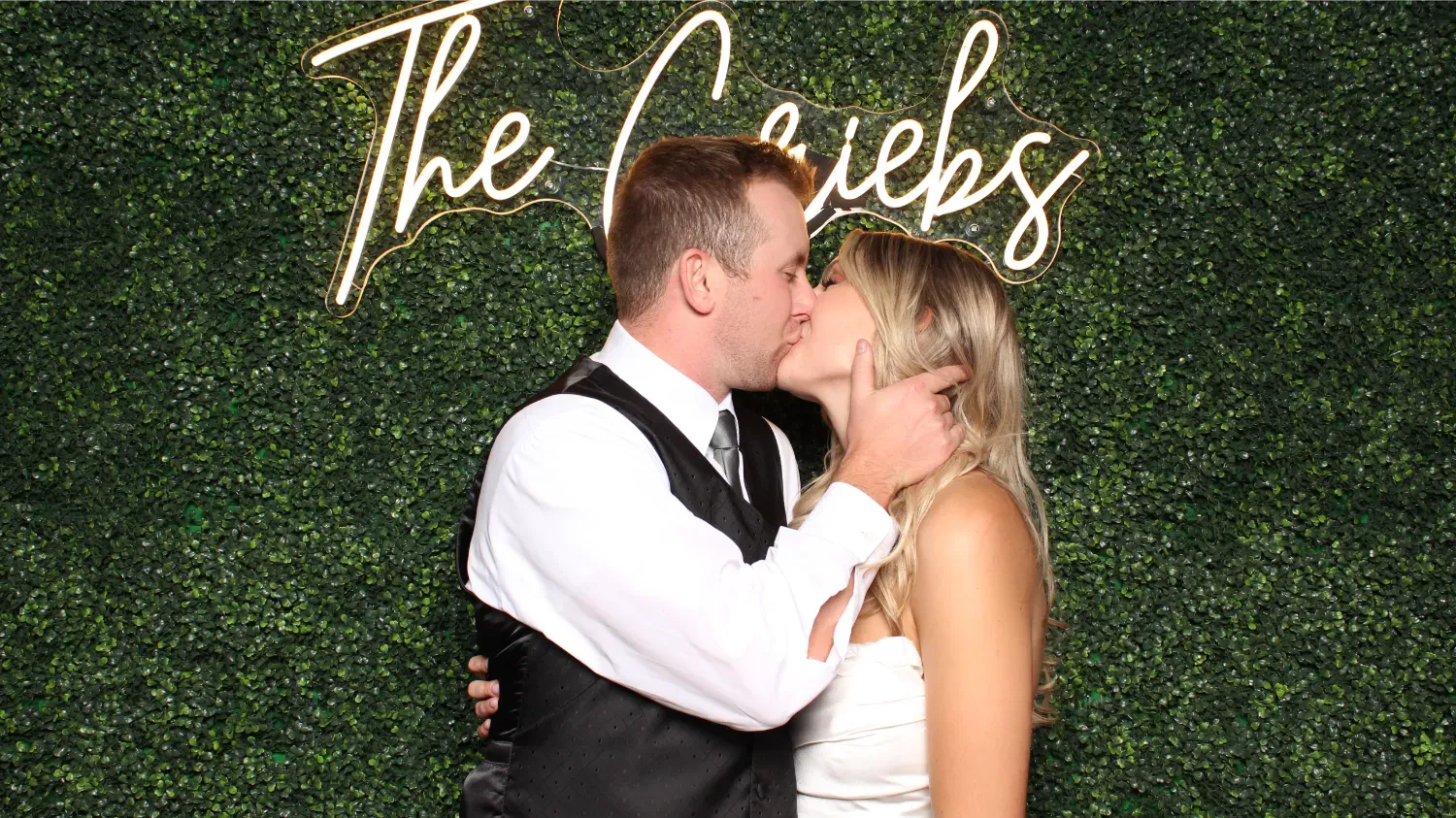 Bride and groom kissing in front of a green hedge wall with a neon sign reading 'The Conciebs'.